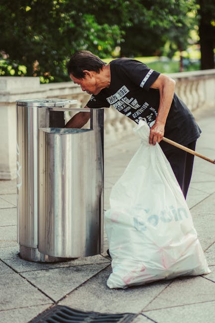 An elderly woman with dark hair pulled back, wearing a black T-shirt with white text and dark pants, is seen bending over a large, cylindrical stainless steel trash bin on a paved outdoor area. She is