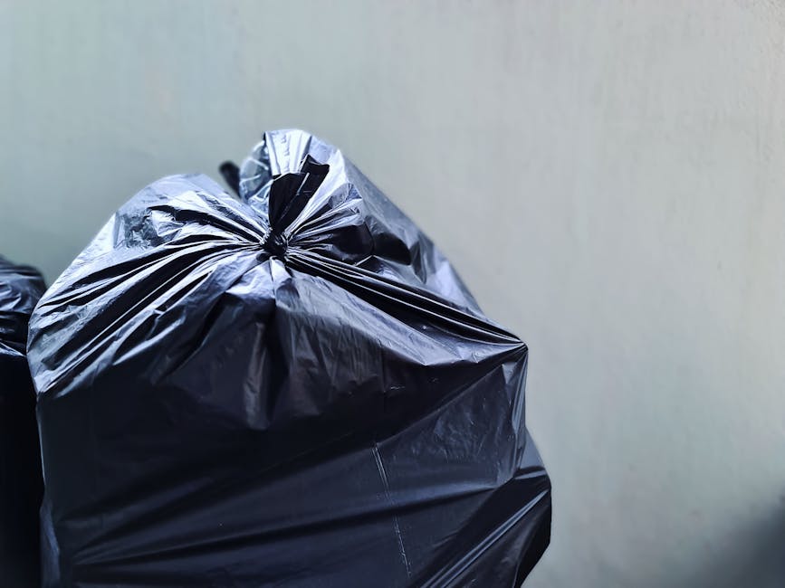 An elderly woman with dark hair pulled back, wearing a black T-shirt with white text and dark pants, is seen bending over a large, cylindrical stainless steel trash bin on a paved outdoor area. She is